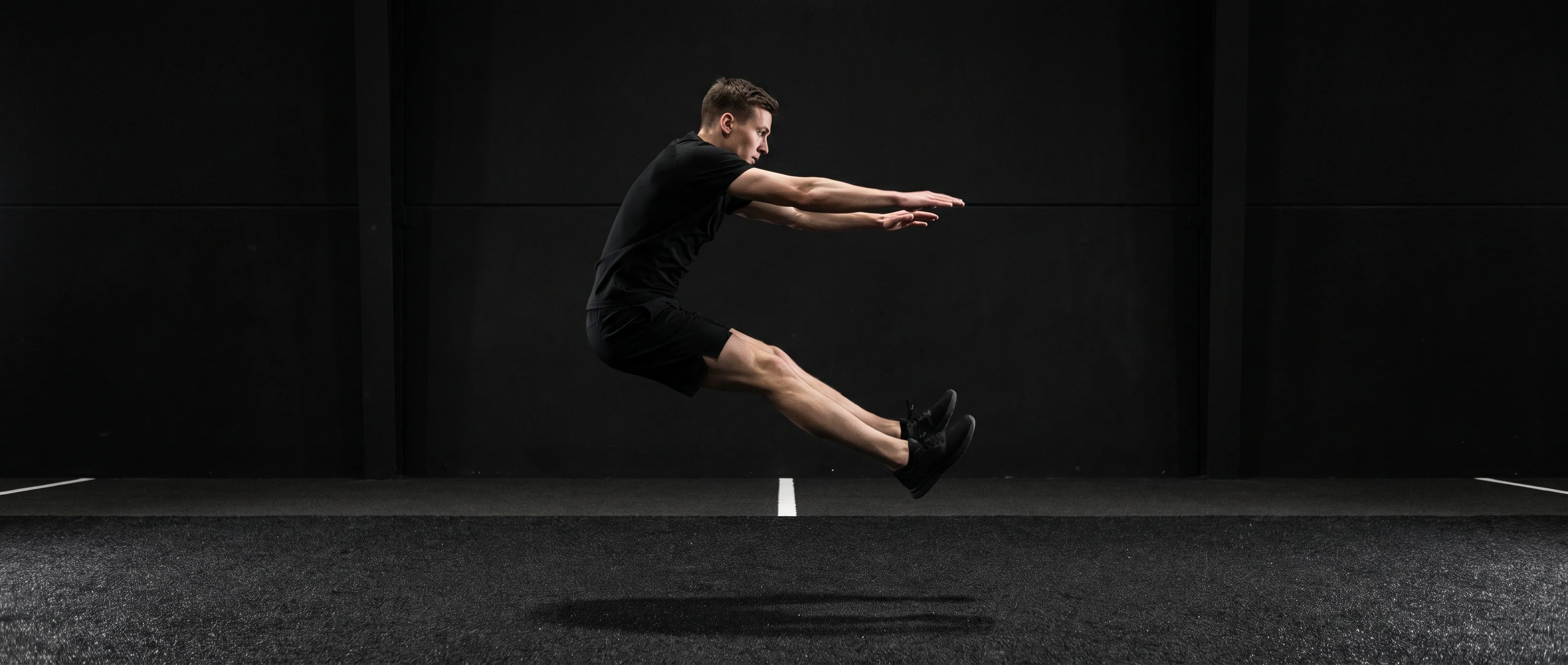 Athlete airborne in a forward two-footed broad jump on dark competition turf