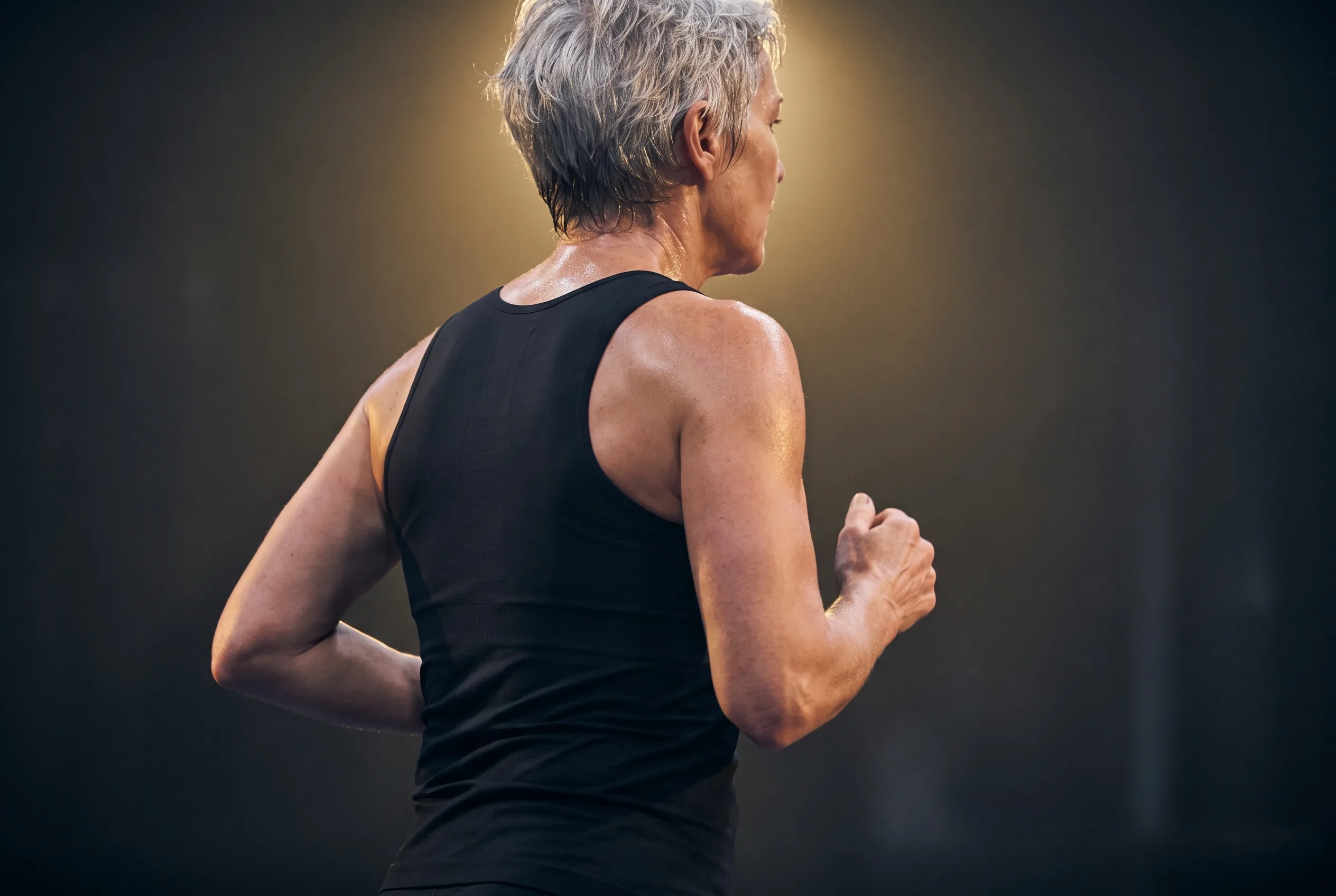 Woman in her sixties running through a dark HYROX arena, rear view