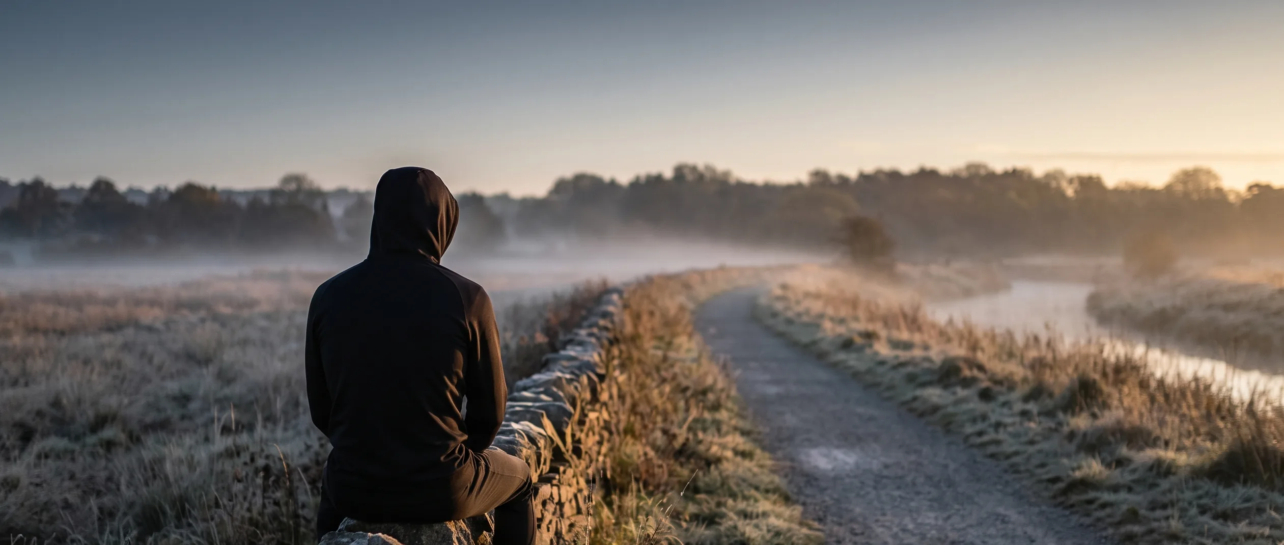 HYROX athlete sitting on a stone wall by a running path at dawn, weighing up overtraining